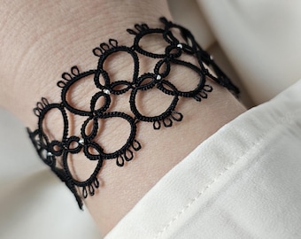Puede incluir: Pulsera de encaje negro con diseño floral. La pulsera se lleva en la muñeca, y el intrincado patrón es visible sobre una camisa blanca. La pulsera parece hecha a mano, con detalles delicados.