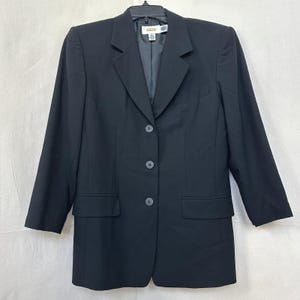 Pode incluir: Um blazer preto liso com lapela entalhada, mangas compridas e fecho frontal de três botões. A peça inclui dois bolsos frontais com aba e uma gola estruturada, com uma etiqueta da marca Talbots visível na parte interna do decote.