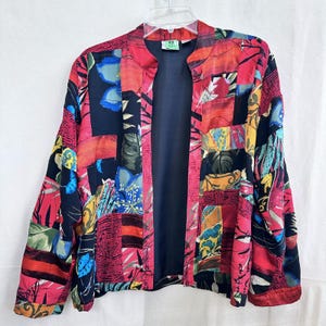 May include: A vibrant, open-front jacket with a patchwork design. The jacket features a mix of red, pink, blue, and yellow floral and abstract patterns. The interior lining is a solid navy blue. The jacket is hanging on a clear hanger.
