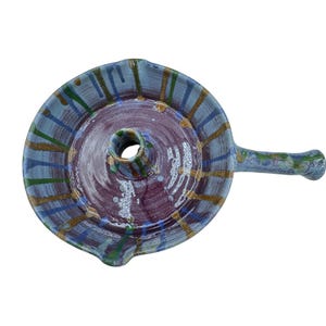 Peut inclure: Lampe à huile en céramique faite à la main avec une poignée. La lampe présente un design circulaire avec un centre violet foncé et un bord bleu clair. Le bord est décoré de rayures vertes, dorées et bleues. La poignée est bleu clair et verte.