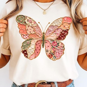 Puede incluir: Camiseta color crema con un gran diseño de mariposa. La mariposa tiene un patrón de patchwork con detalles florales, cuadros y encaje en tonos rosa, verde y rojo, contorneados en dorado. La camiseta se combina con vaqueros azules y un cinturón marrón.
