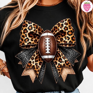 Op de afbeelding: Een zwart t-shirt met een grote afbeelding van een bruine football in het midden van een decoratieve strik. De strik toont een mix van luipaardprints, zwarte glittersecties en gouden glitteraccenten, wat zorgt voor een opvallend visueel ontwerp.