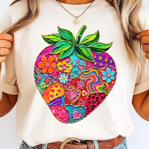 Puede incluir: Una camiseta color crema con un diseño de fresa vibrante y colorido. La fresa está compuesta por varios patrones, incluyendo flores, corazones y espirales, en tonos de rosa, azul, verde y amarillo. La camiseta se combina con vaqueros azules y un cinturón marrón.