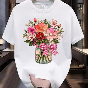 Puede incluir: Camiseta blanca con un ramo floral colorido en un tarro de cristal. El ramo incluye flores rojas, rosas y melocotón con hojas verdes. Un lazo rosa y blanco a cuadros adorna el tarro. La camiseta tiene cuello redondo y mangas cortas.