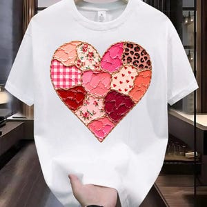 Puede incluir: Camiseta blanca con un diseño en forma de corazón compuesto por varios patrones de tela. El corazón incluye parches florales, de cuadros y de leopardo en tonos rosa, rojo y melocotón, con un borde dorado. La camiseta es sostenida por una mano.