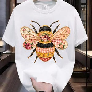 Puede incluir: Camiseta blanca con un diseño de abeja grande y colorido. La abeja tiene un cuerpo a rayas negro y amarillo, detalles florales y alas estampadas con rosas rosas y rojas. Prenda casual.