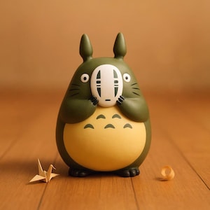 Puede incluir: Una figura verde y amarilla de un personaje de dibujos animados que sostiene una máscara blanca con detalles negros. La figura tiene un cuerpo redondo, orejas puntiagudas y ojos negros. Una grulla de papel y un pequeño trozo de papel enrollado están al lado.