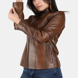 Könnte beinhalten: Braune Lederjacke mit Stehkragen und Reißverschluss. Die Jacke hat eine taillierte Silhouette und verfügt über vertikale Nähte und Taschen. Das Model trägt eine blaue Jeans.