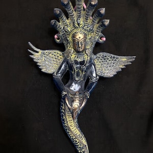 May include: A detailed, blue and gold statue of a deity with multiple cobra heads, wings, and a serpent-like lower body. The statue has intricate detailing and is set against a black background. The figure is holding a small object.