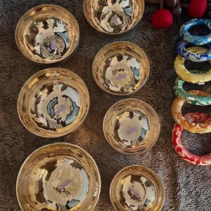 May include: A collection of seven golden singing bowls with a hammered texture, arranged on a textured surface. The bowls vary in size. Also visible are colorful patterned bracelets and red felt mallets.