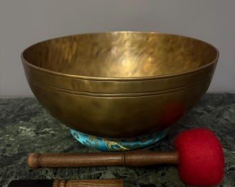 Peut inclure: Un grand bol chantant doré avec un maillet en bois et un petit percuteur en bois. Le bol repose sur un coussin bleu et or. Le maillet a une tête en feutre rouge.