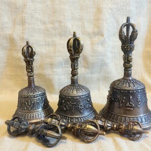 Op de afbeelding: Drie antieke bronzen bellen met sierlijke details en decoratieve handvatten. De bellen zijn van verschillende formaten en worden vergezeld door drie bronzen vajra's. De bellen en vajra's hebben ingewikkelde patronen en ontwerpen.