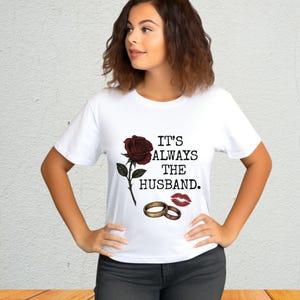 Op de afbeelding: Wit T-shirt met de tekst "IT'S ALWAYS THE HUSBAND." met een rode roos, twee gouden trouwringen en een rode lipafdruk. Het shirt is een casual stijl.