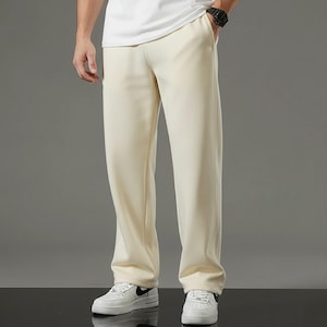 Op de afbeelding: Een persoon draagt een wit T-shirt met ronde hals en een crèmekleurige joggingbroek. De persoon draagt ook een zwart horloge en witte sneakers. De outfit is casual en comfortabel.