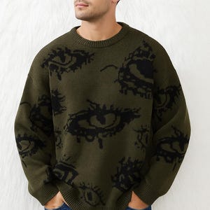 Puede incluir: Suéter verde oliva con cuello redondo y un patrón de diseños de ojos negros. El suéter es de punto y tiene mangas largas. Una prenda casual y cómoda.