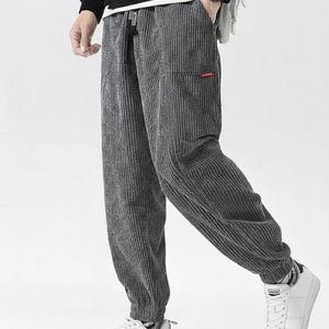 May include: Gray corduroy pants with a drawstring waist and a small red accent on the pocket. The pants have a relaxed fit and are paired with white sneakers. The corduroy fabric has vertical ridges.