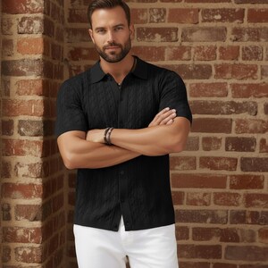 May include: Black short-sleeve knit shirt with a textured pattern and a collared neckline. The shirt is buttoned down the front and is paired with white pants. The background is a brick wall.