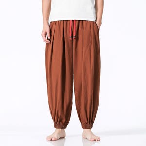 May include: Brown harem pants with a red drawstring. The pants have a loose fit with gathered ankles. The person in the image is wearing a white t-shirt. The pants are made of a soft material.