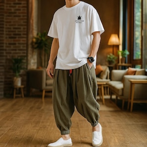 May include: A person wearing a white t-shirt with the text "AMERICAN ANARS" and a small graphic. They are also wearing olive green harem pants and white slip-on shoes. A black watch is on the person's wrist.