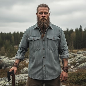 May include: A man wearing a green striped shirt with two chest pockets, standing outdoors. He is holding an axe and has a long beard. The shirt has a button-down design and is made of a striped fabric. The background features a forest and a cloudy sky.