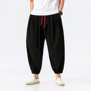 May include: Black harem pants with a red drawstring. The pants have a loose fit with gathered ankles. The person is wearing a white t-shirt and white sneakers.