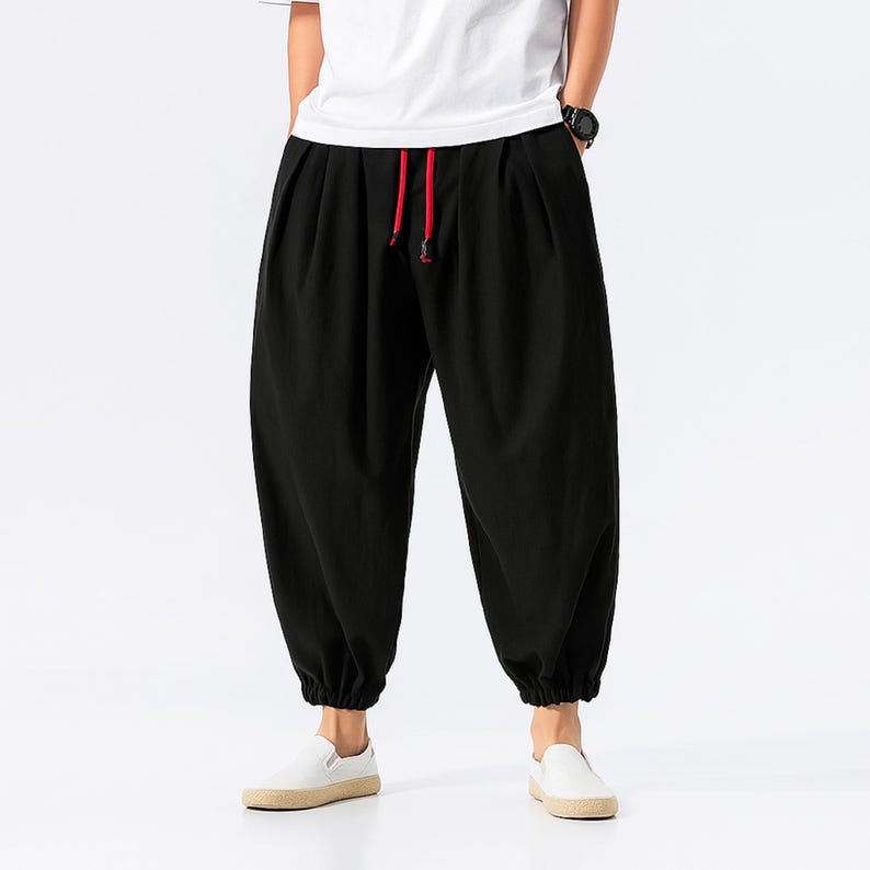 May include: Black harem pants with a red drawstring. The pants have a loose fit with gathered ankles. The person is wearing a white t-shirt and white sneakers.