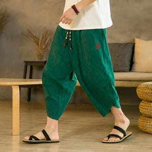 May include: Green, loose-fitting pants with a textured pattern and drawstring waist. The pants are paired with black sandals. A white top is partially visible. The pants have a small red square patch.