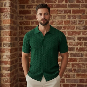 Puede incluir: Un polo verde oscuro de manga corta con un patrón de punto trenzado. La camisa tiene una botonadura y un cuello clásico. La persona lleva la camisa con pantalones claros, de pie frente a una pared de ladrillos.