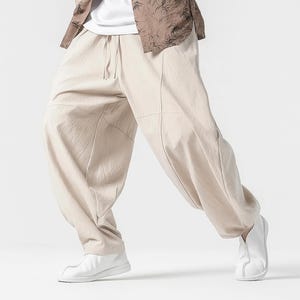 Op de afbeelding: Beige, loszittende broek met een tailleband met trekkoord en gestikte paneeldetails. De broek wordt gecombineerd met witte schoenen en een overhemd met knopen en een patroon. De stijl suggereert een casual, comfortabele uitstraling.