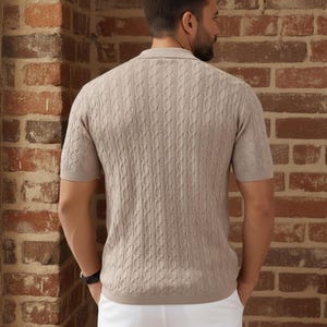May include: A light brown short-sleeve polo shirt with a cable knit pattern. The shirt has a collared neckline and is paired with white pants. The background is a brick wall.