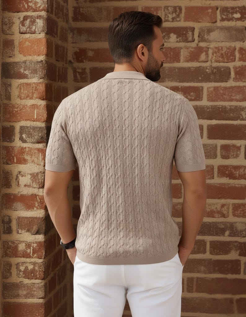 May include: A light brown short-sleeve polo shirt with a cable knit pattern. The shirt has a collared neckline and is paired with white pants. The background is a brick wall.