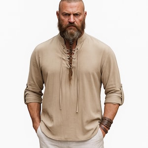 May include: Tan long-sleeve shirt with a lace-up neckline and rolled-up sleeves. The shirt has a mandarin collar and is made of a lightweight fabric. The model is wearing a brown leather bracelet. The shirt is a casual style.