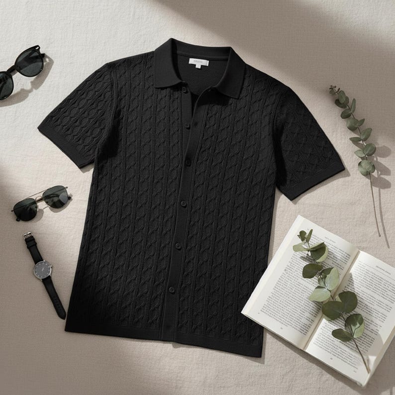May include: Black short-sleeve polo shirt with a textured cable knit pattern. The shirt has a button-down front and a classic collar. Sunglasses, a watch, and an open book are also visible.