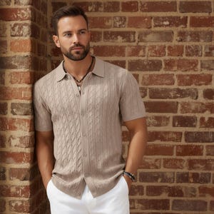 May include: A light brown short-sleeve shirt with a cable knit design. The shirt has a button-up front and a collared neckline. The person is wearing white pants and a beaded necklace.