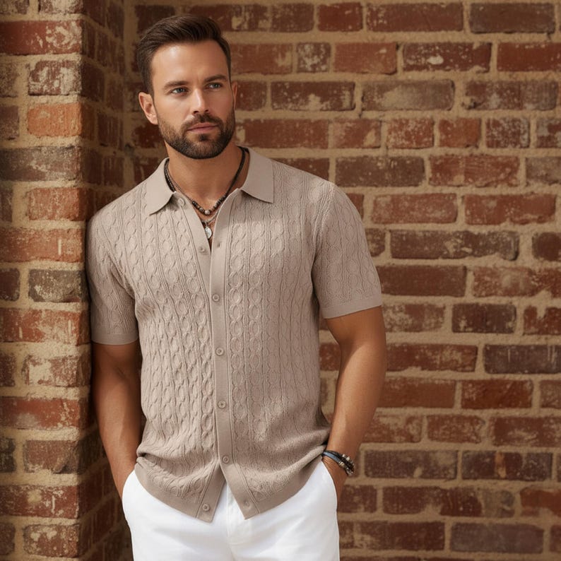 May include: A light brown short-sleeve shirt with a cable knit design. The shirt has a button-up front and a collared neckline. The person is wearing white pants and a beaded necklace.