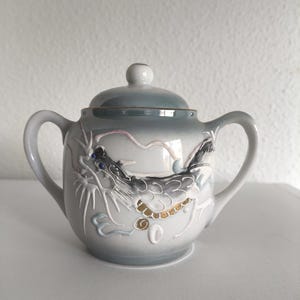 Peut inclure: Un sucrier en céramique gris avec un couvercle, orné d'un motif de dragon. Le bol présente un effet dégradé, et le dragon est représenté en noir, or et bleu. Il possède deux anses et un petit bouton sur le couvercle.