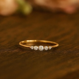 Puede incluir: Un delicado anillo de oro con cinco piedras preciosas redondas y transparentes dispuestas en fila. La banda del anillo es fina y pulida, y las piedras preciosas brillan. El anillo se muestra sobre una superficie de madera oscura.