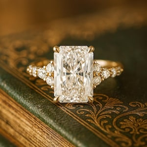 May include: A radiant-cut diamond engagement ring with a gold band. The ring features a large, rectangular diamond as the centerpiece, flanked by smaller accent diamonds. The ring is set on a vintage book with gold-embossed detailing.