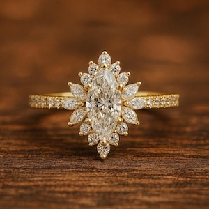 Puede incluir: Anillo de compromiso dorado con una piedra central de diamante talla marquesa, rodeada de diamantes más pequeños en un diseño de halo floral. La banda también está adornada con pequeños diamantes, sobre un fondo de madera.