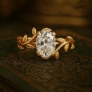 Pode incluir: Anel de ouro com um diamante central de corte oval. A banda do anel é adornada com detalhes delicados de folhas e videiras, criando um design elegante inspirado na natureza. O anel está sobre um fundo escuro com padrões.