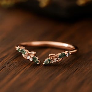 Puede incluir: Anillo de oro rosa con diseño de hojas. El anillo presenta piedras preciosas verdes y pequeñas gemas blancas. El anillo está sobre una superficie de madera.