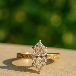 Puede incluir: Un anillo de compromiso con un diamante talla marquesa y una banda de oro. El diamante es el punto focal, engastado en un engaste de cuatro puntas. La banda de oro presenta un diseño intrincado y grabado. El anillo se muestra sobre una superficie neutra con un fondo verde borroso.