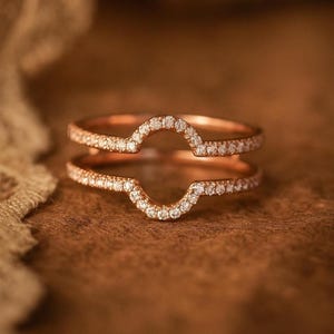 Può includere: Anello in oro rosa con un design curvo, incastonato con piccole pietre preziose bianche scintillanti. L'anello ha due fasce, ciascuna bordata dalle pietre preziose, creando un effetto alone. L'anello è posto su uno sfondo marrone caldo.