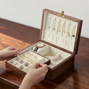 Puede incluir: Una caja de joyería de madera con la tapa abierta, que revela compartimentos llenos de joyas. La caja es marrón con un interior de color claro. Se ven un reloj y varios collares, pulseras y anillos.