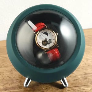 May include: A green watch display case with a clear, rounded front. Inside, a gold-toned watch with a white face, moon phase indicator, and red leather band is visible. The case has silver-colored feet.