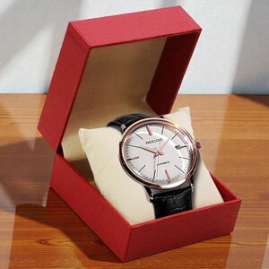 May include: A wristwatch with a white face, rose gold accents, and a black leather band is displayed in an open red gift box. The watch face has the brand name "ROCOS" and the word "AUTOMATIC" printed on it. The watch is resting on a cream-colored pillow.