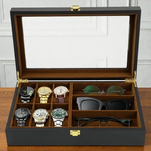 Puede incluir: Una caja de exhibición de madera negra para relojes y gafas con tapa de cristal. El interior está forrado con terciopelo marrón y contiene varios relojes y pares de gafas. La caja tiene un cierre y bisagras doradas.