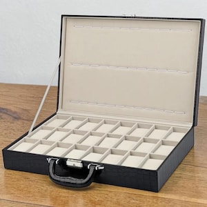 May include: Open black faux leather watch case with a handle and silver-tone clasp. The interior features a beige lining and individual compartments for watch storage. The case is open, revealing the compartments.