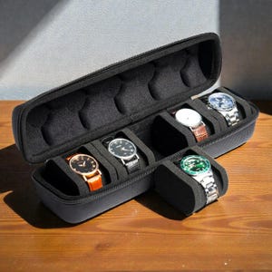 May include: Black travel watch case with six compartments, each holding a different wristwatch. The watches have various band colors, including brown, orange, and silver. The case is open, revealing the watches inside.