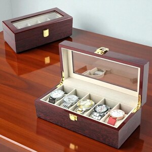 May include: A wooden watch box with a glass top, showcasing five wristwatches. The box is a rich, dark brown with a gold-colored clasp. The interior is lined with a light beige fabric. A second, smaller watch box is visible in the background.